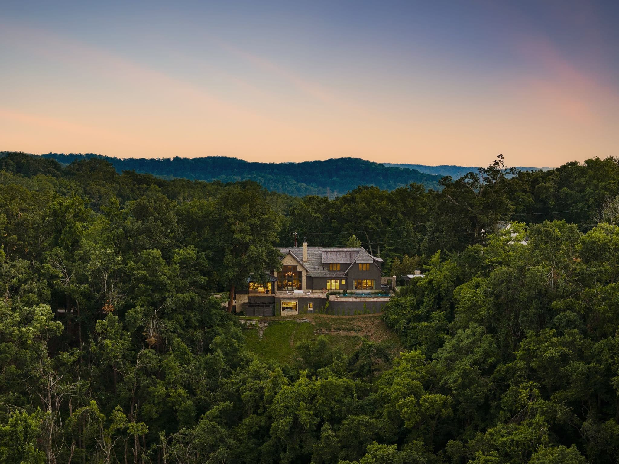 Elevated dusk view of a contemporary hillside estate at 1801 Laurel Ridge Drive in Forest Hills, Nashville, with lit interiors, a pool terrace and a wooded ridgeline beyond